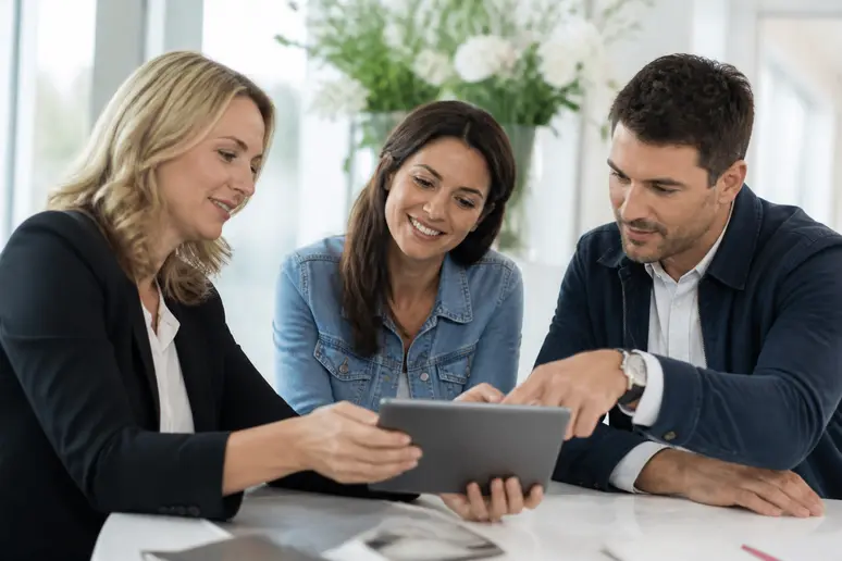 Une organisatrice de mariage présente des options de lieux de réception à un couple sur une tablette dans un bureau lumineux.