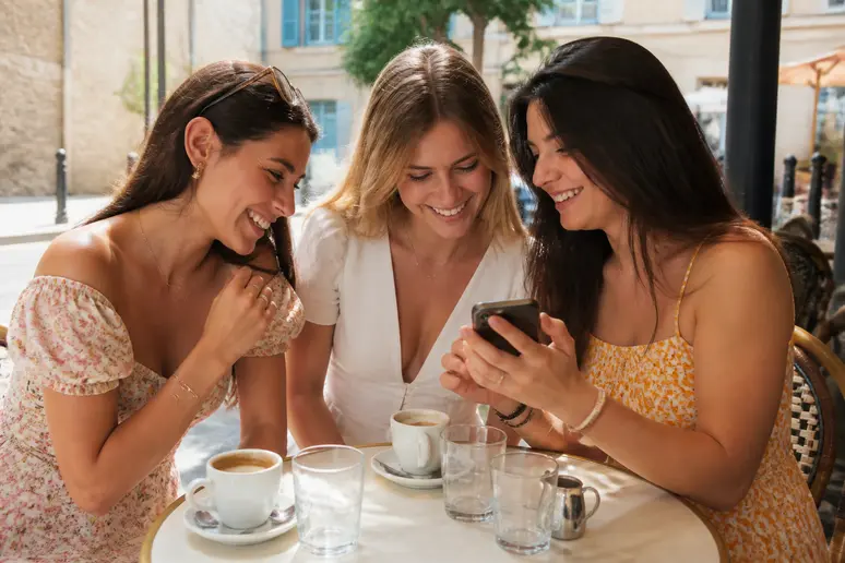 Trois amies complices trient des photos de mariage sur un smartphone, dans une ambiance décontractée et ensoleillée.