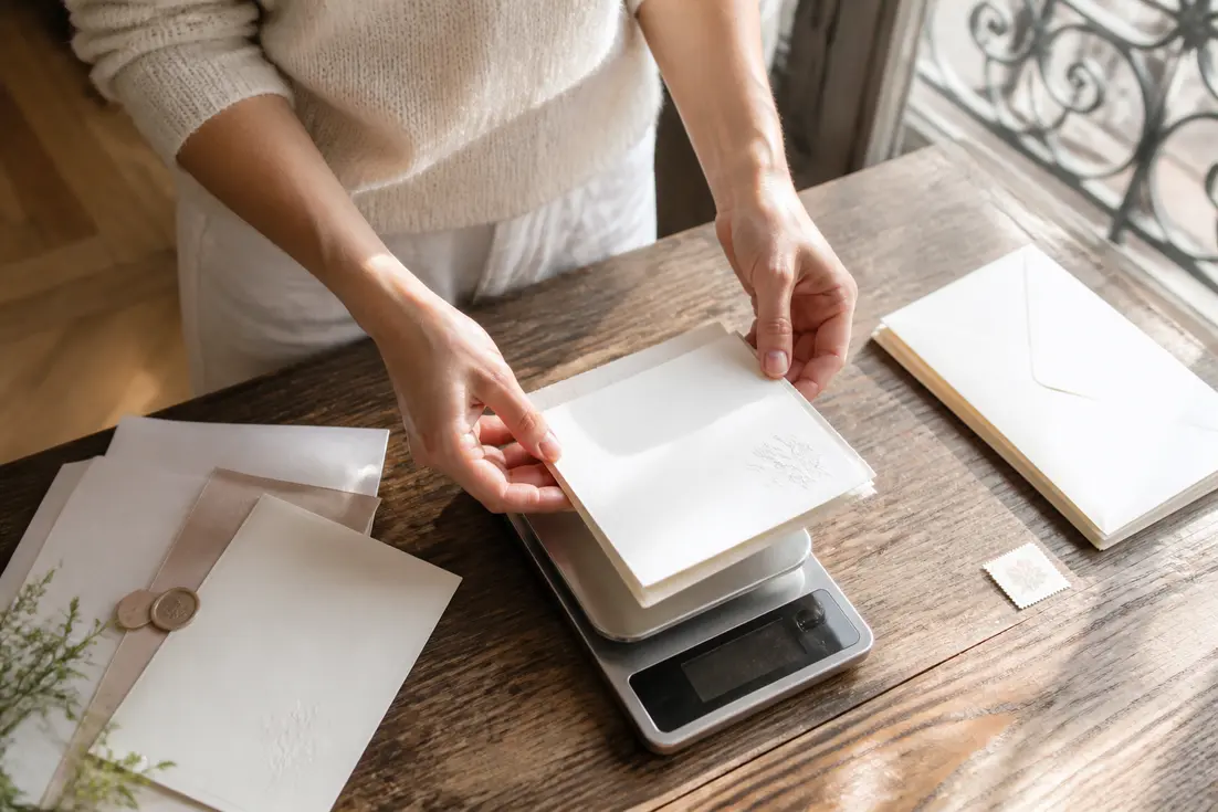 Mains pesant un faire-part sur une balance pour savoir comment choisir enveloppe mariage sans dépasser vingt grammes