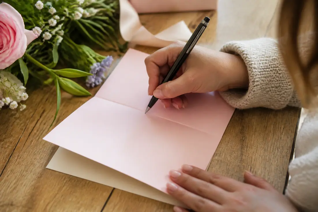 Une personne écrit un texte félicitation naissance fille sur une jolie carte rose pastel